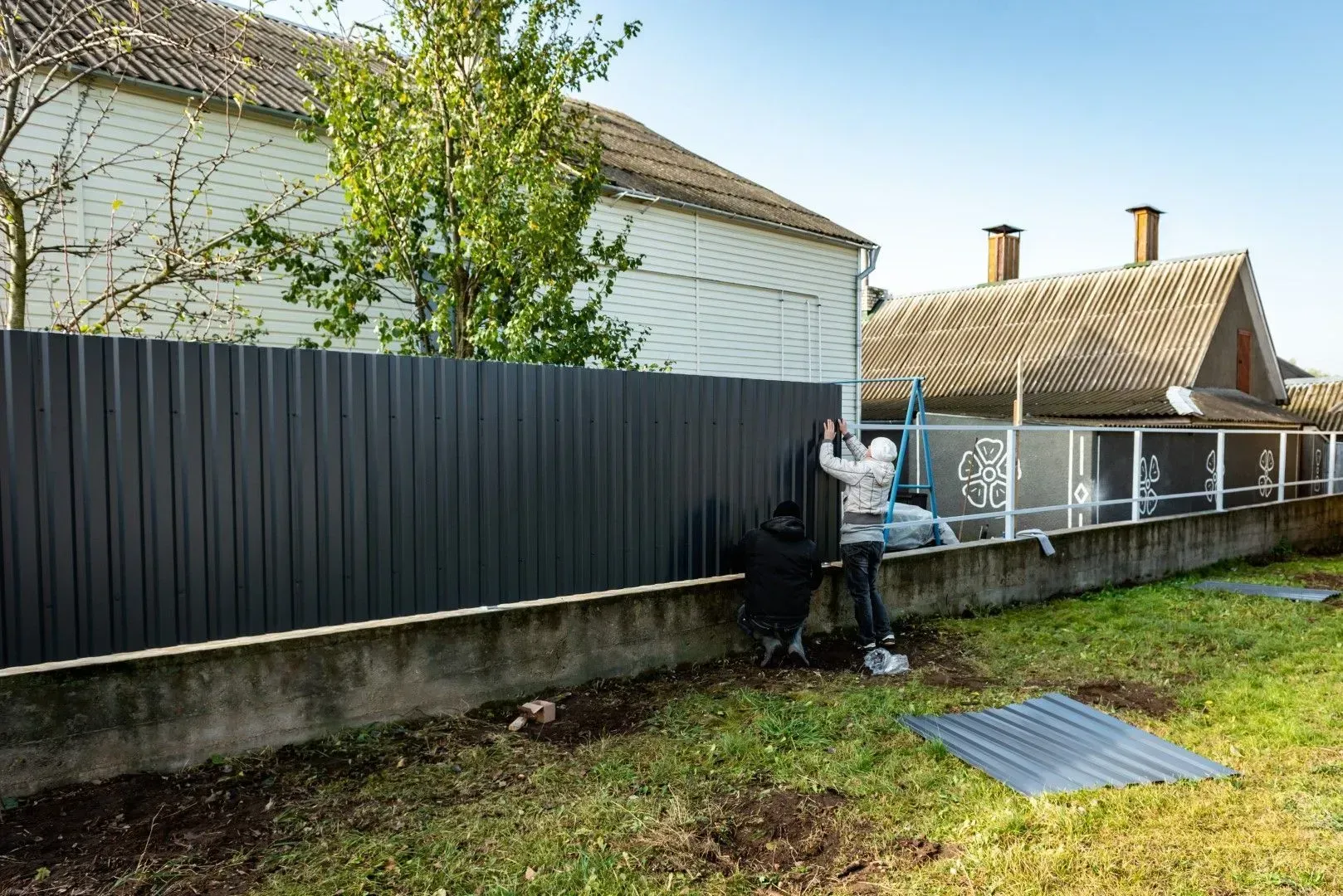 Person working by a black fence in a grassy backyard beside a house, with a metal grate on the ground
