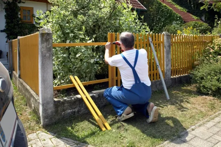 Person in white shirt and blue overalls attaching a wooden fence rail in a garden