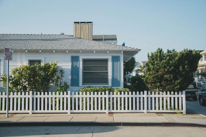 Small white house with blue shutters and a white picket fence on a quiet street.