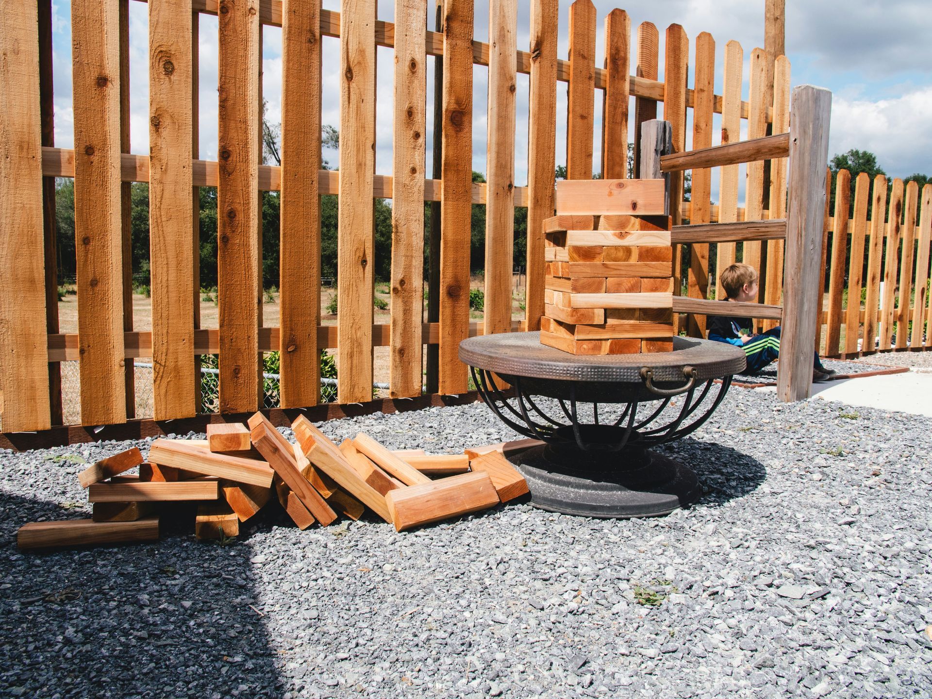 Rustic outdoor fire pit area with stacked firewood beside a wooden fence on gravel.