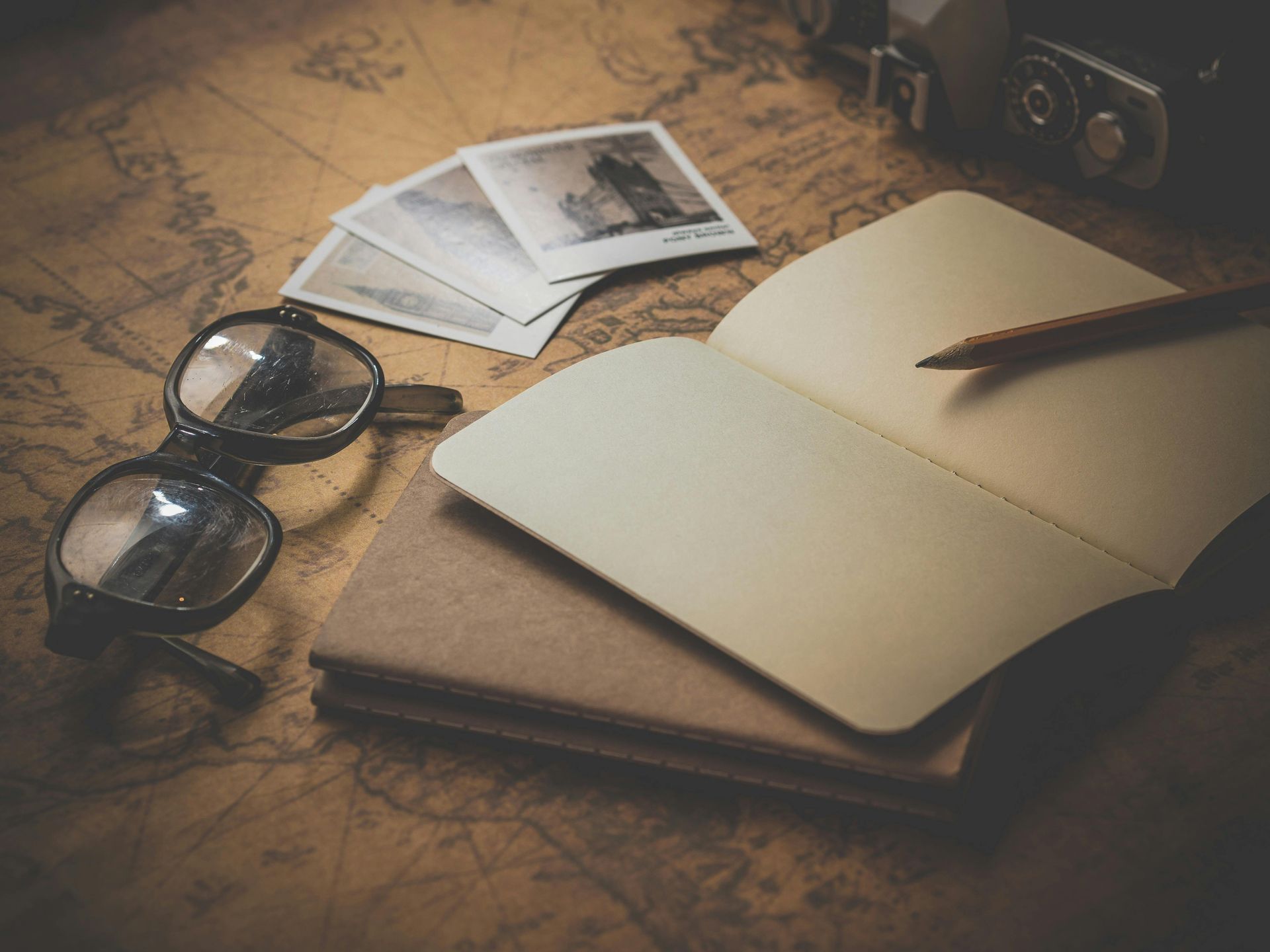 Open notebook and glasses on a world map, with photos and camera, implying travel and writing.