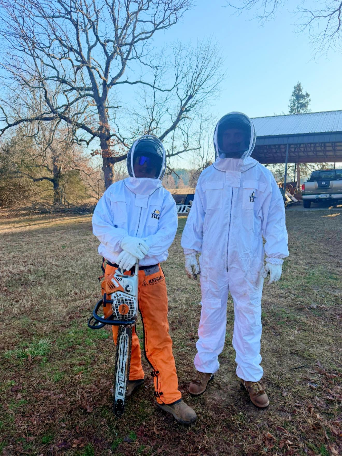 Two children in white protective suits and helmets stand outdoors beside a tree and an orange chainsaw.
