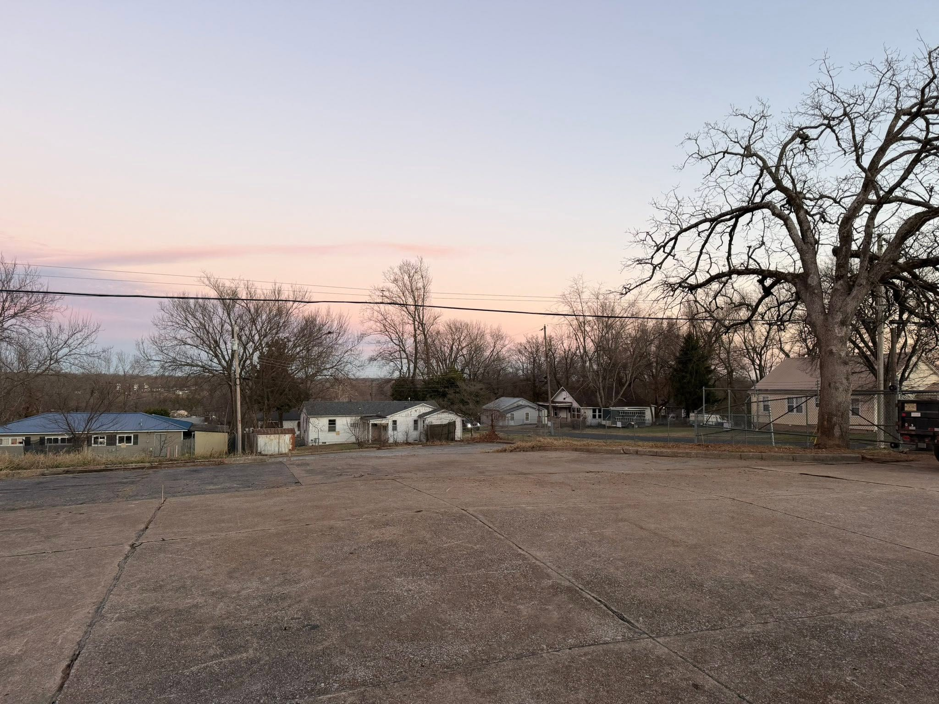 Empty gravel lot at dusk with bare trees, small houses, and pink sky in the background