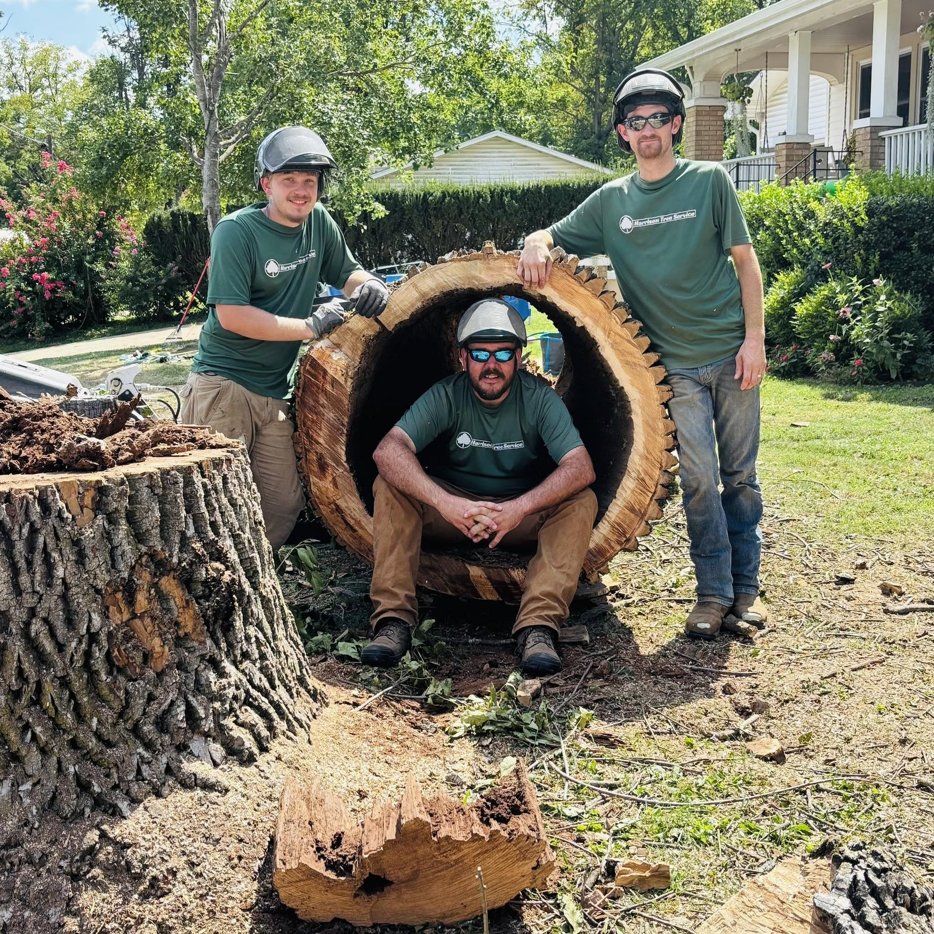 Three workers in green shirts pose with a huge cut log section in a yard.