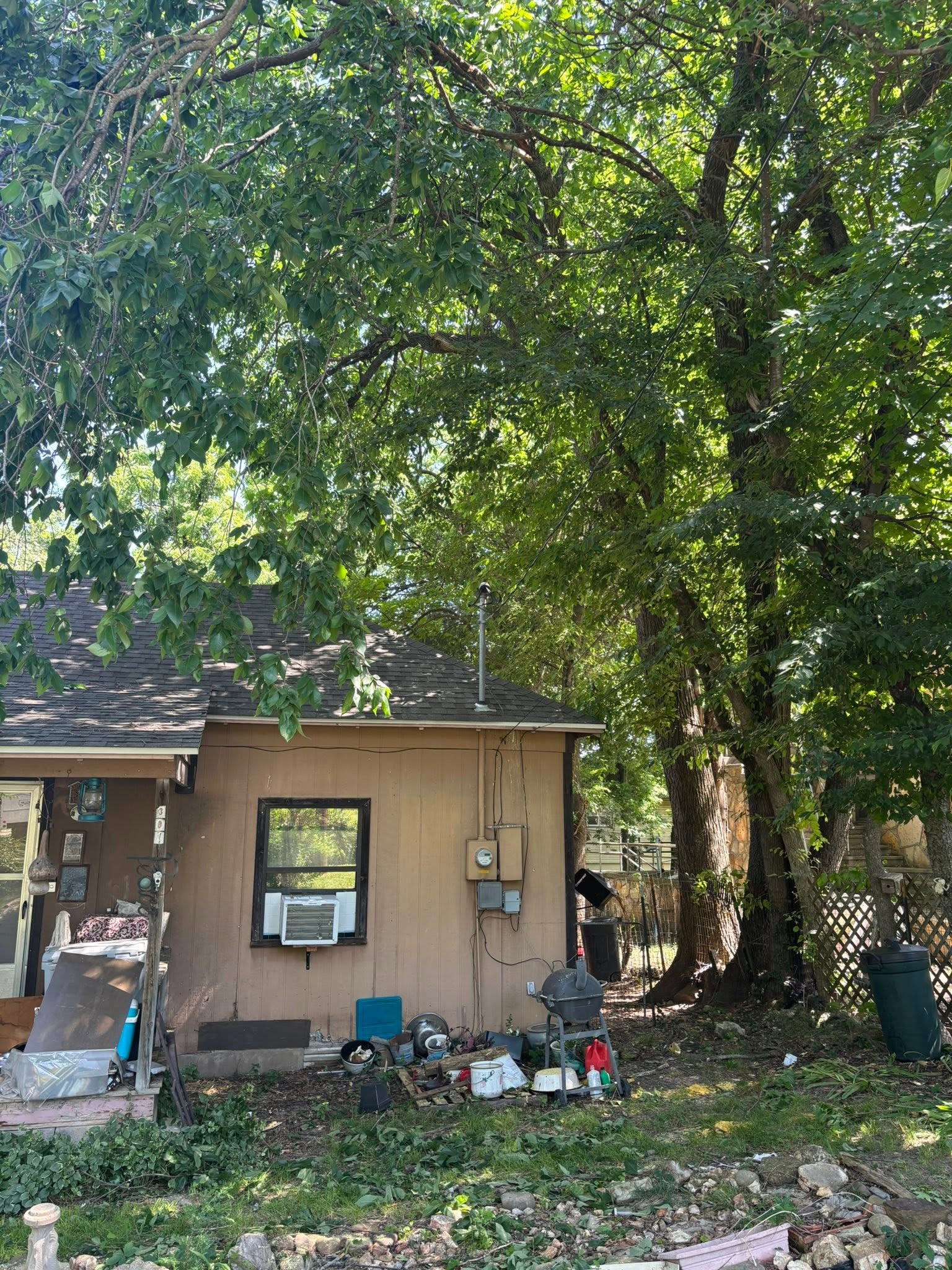 Small tan house beneath large leafy trees, with a shaded yard and scattered outdoor items.