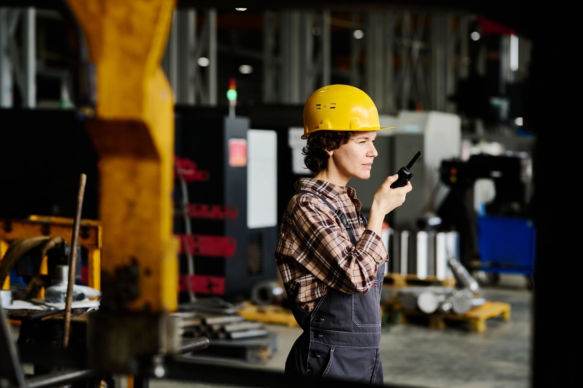 Worker wearing a hard hat and coveralls using a two-way radio inside an industrial facility.