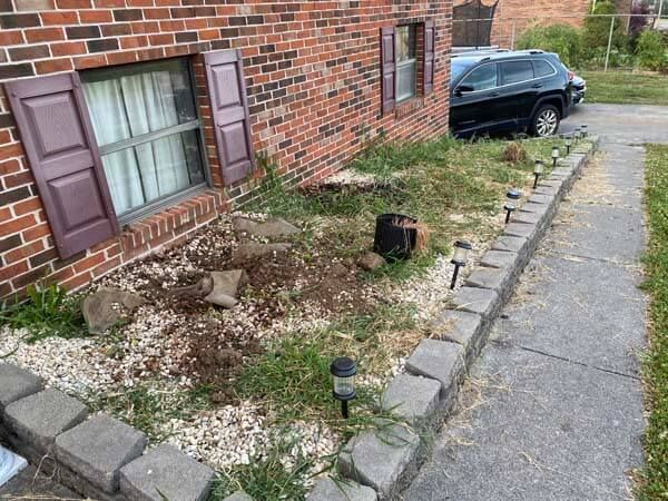 Small brick house garden bed with rocks, sparse plants, and a sidewalk beside a parked car