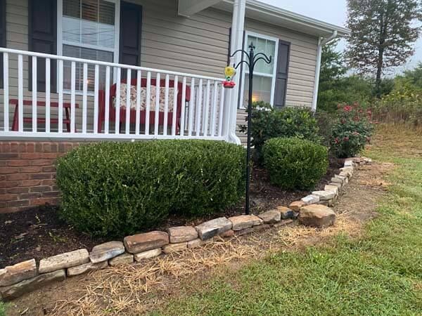 Front porch with white railing, trimmed shrubs, and a stone border along the house foundation.