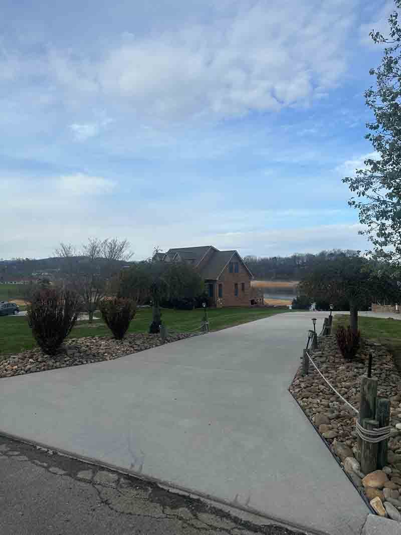 Long driveway leading to a brick house, with shrubs, stone edging, and cloudy sky above.