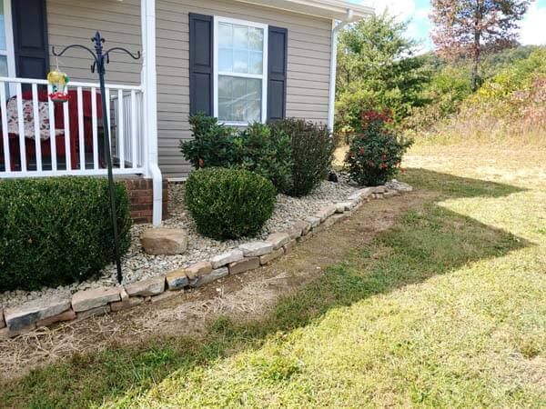 Front yard landscaping with shrubs, stone edging, and a house porch with white railing