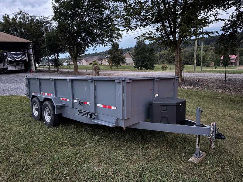 Gray dual-axle utility trailer with hitch parked on grass near trees and a gravel area