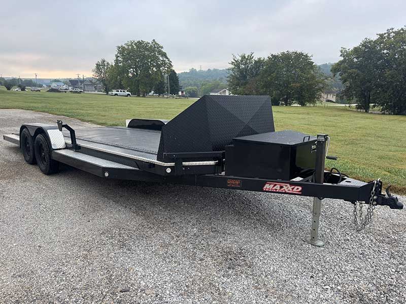 Black flatbed utility trailer parked on gravel beside a grassy field, with a diamond-plate toolbox.