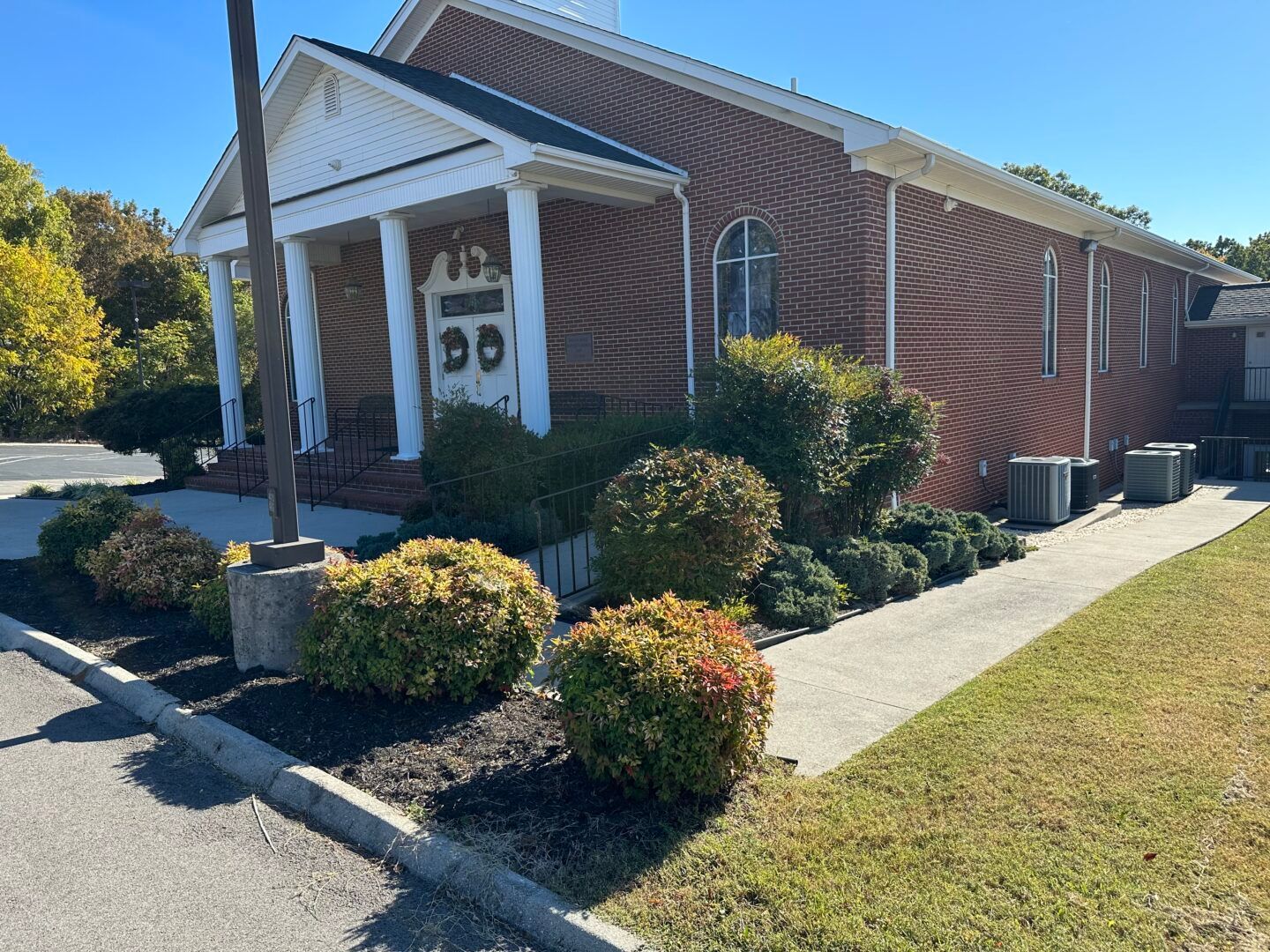 Brick building with white columns, shrubs, and a sidewalk on a sunny day