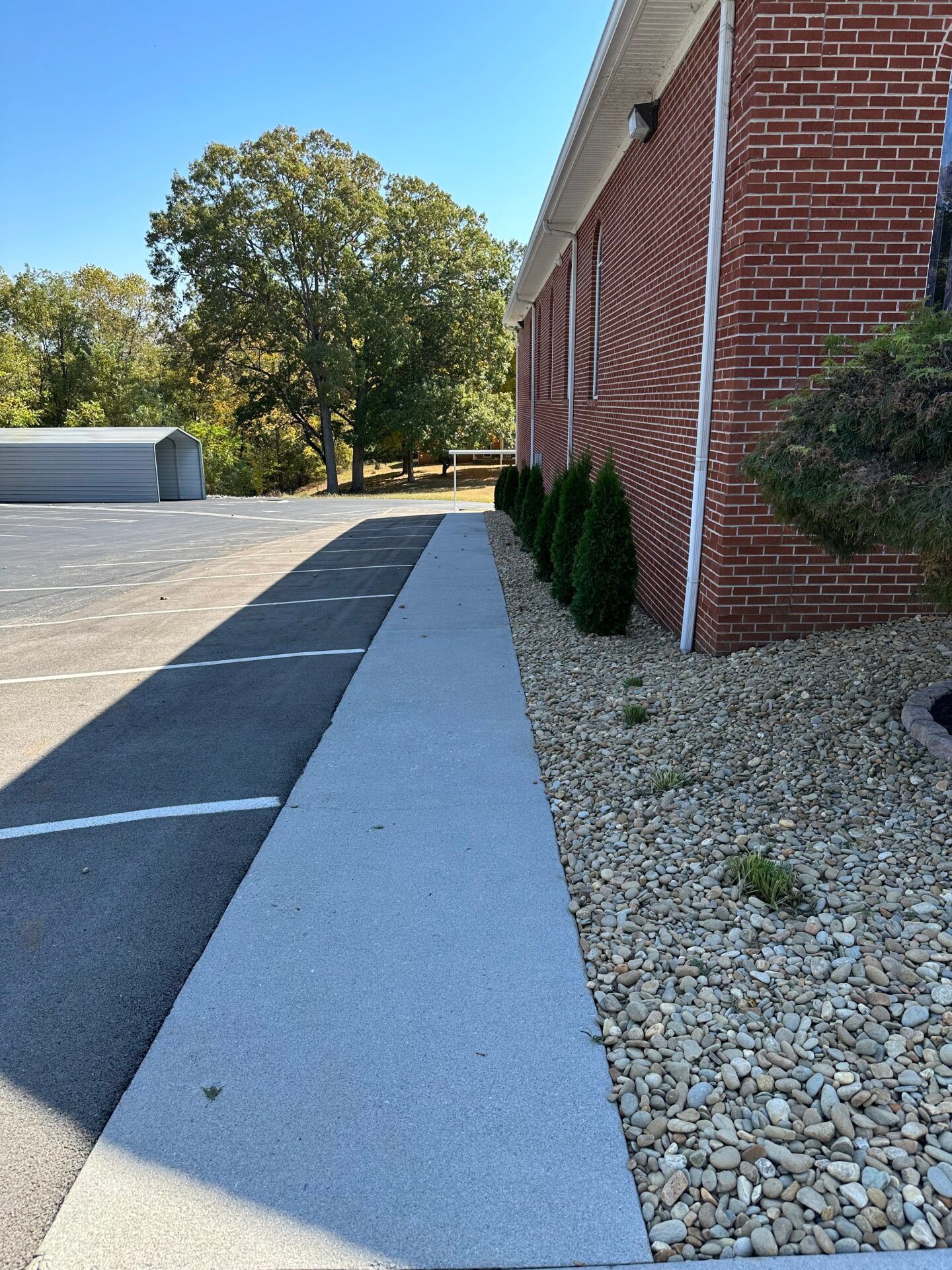 Parking lot beside red brick building with white sidewalk, gravel border, and trees in the background
