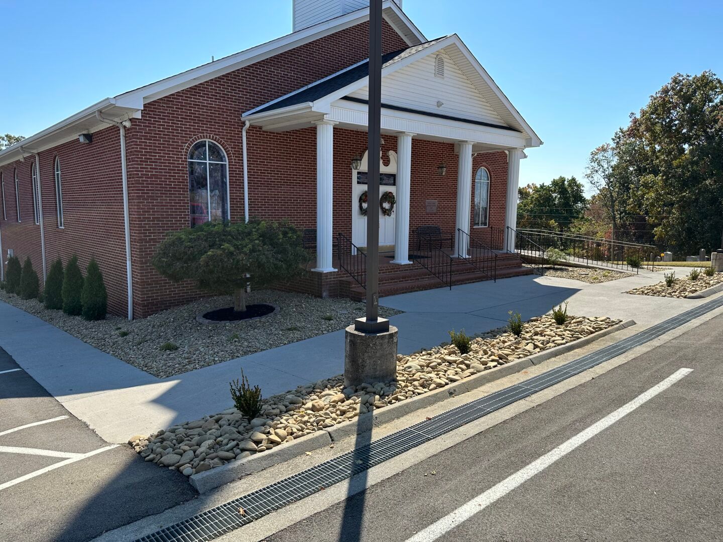 Brick church entrance with white columns, ramp, and roadside gravel landscaping under a clear sky