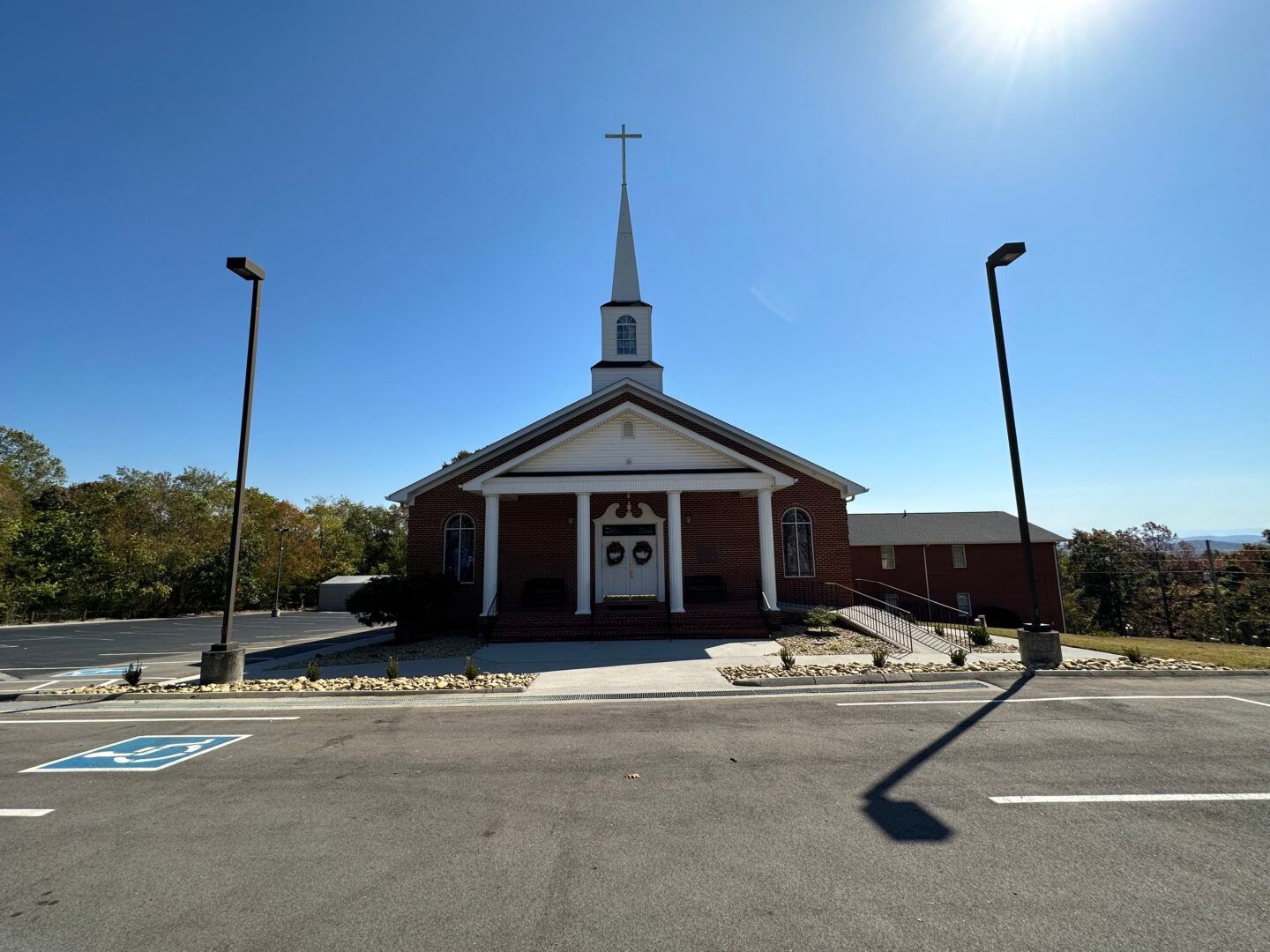 Small white church with a steeple and cross, viewed from across a parking lot on a sunny day.