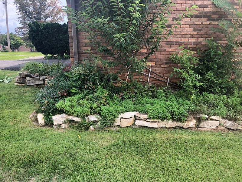 Brick house corner with a small rock-edged garden bed and green shrubs beside a lawn