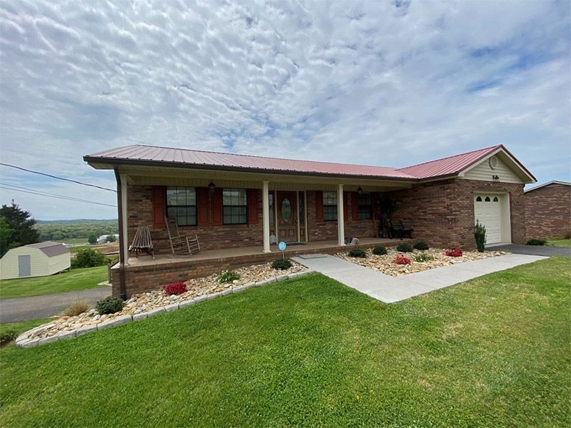 Single-story brick house with red roof, front porch, and landscaped lawn under a cloudy sky