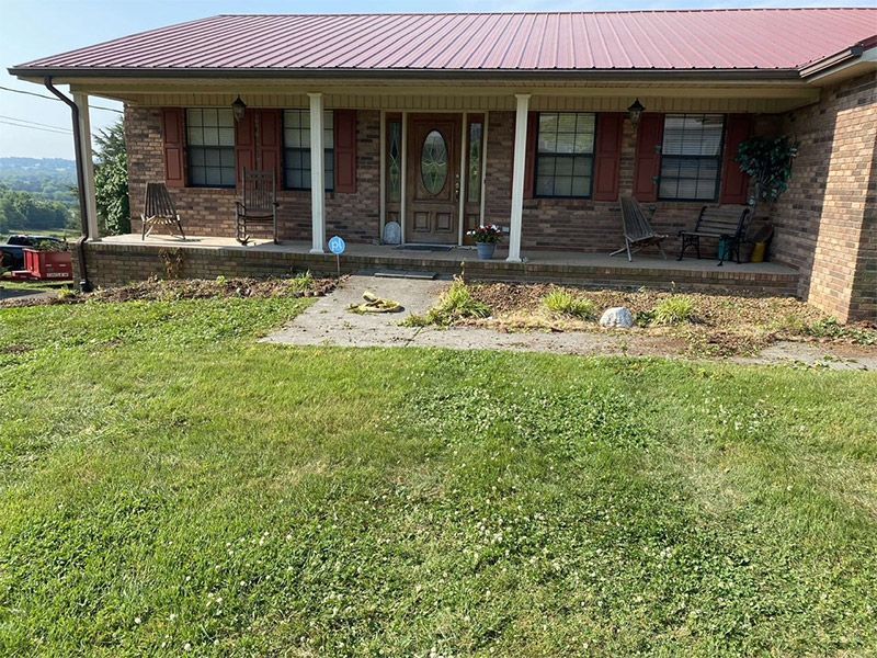 Brick house with red metal roof and front porch, set behind a grassy yard.