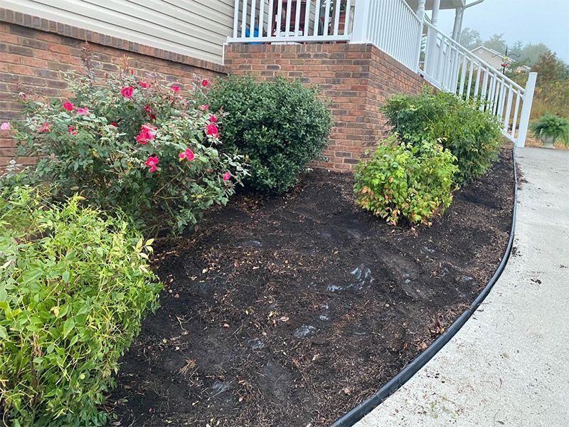 Curved garden bed with shrubs and pink flowers beside a brick house and sidewalk