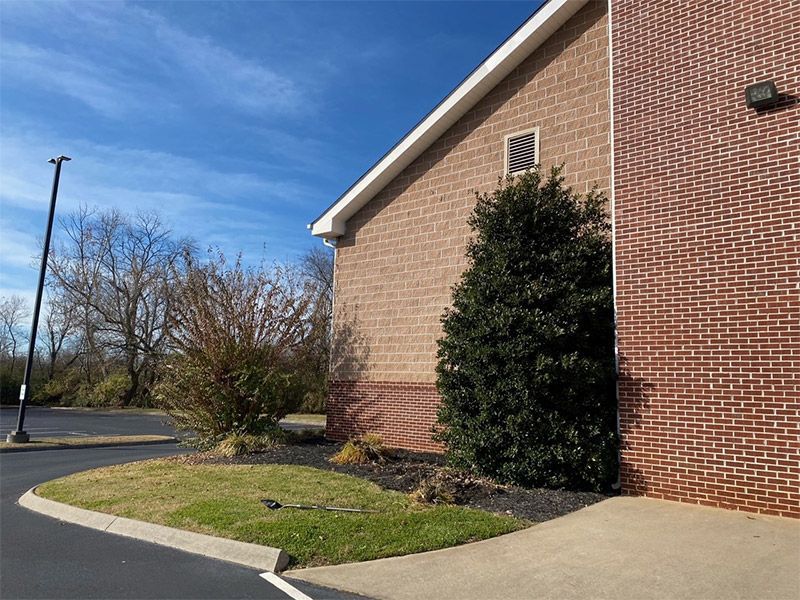 Brick building corner with shrubs, grass, and a curved sidewalk under a blue sky.