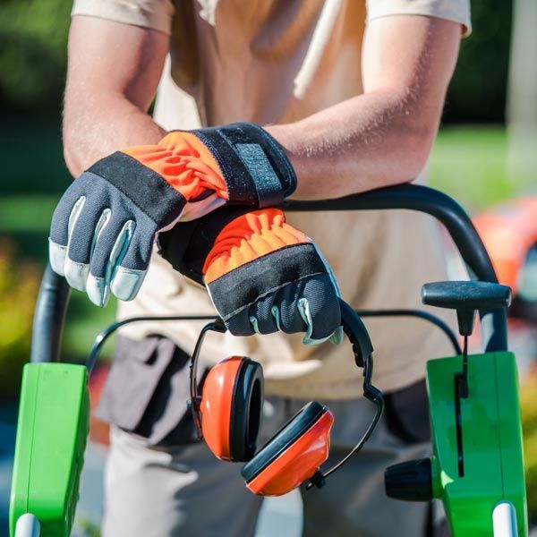 Gloved worker leaning on a wheelbarrow with orange earmuffs hanging from it outdoors