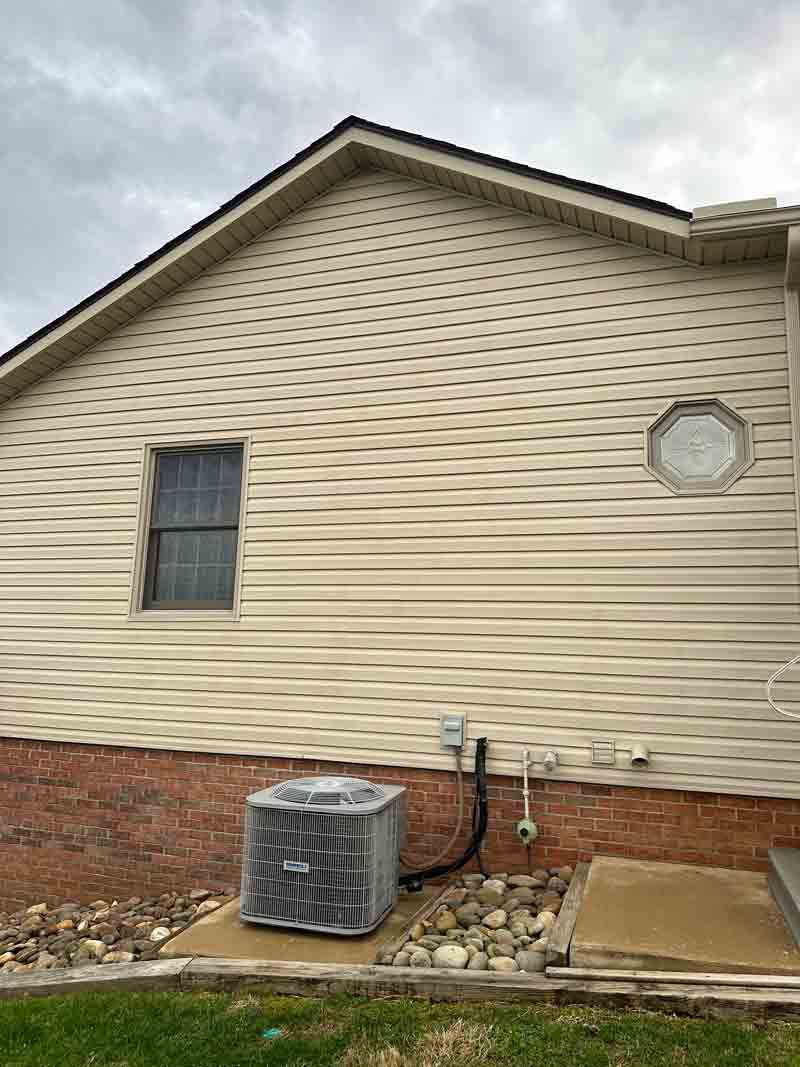Side of a beige house with a window, round attic vent, and air conditioner unit by the foundation.