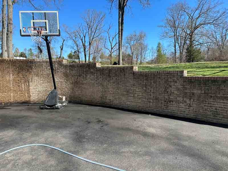 Outdoor basketball hoop beside a brick wall on a paved court under a clear blue sky