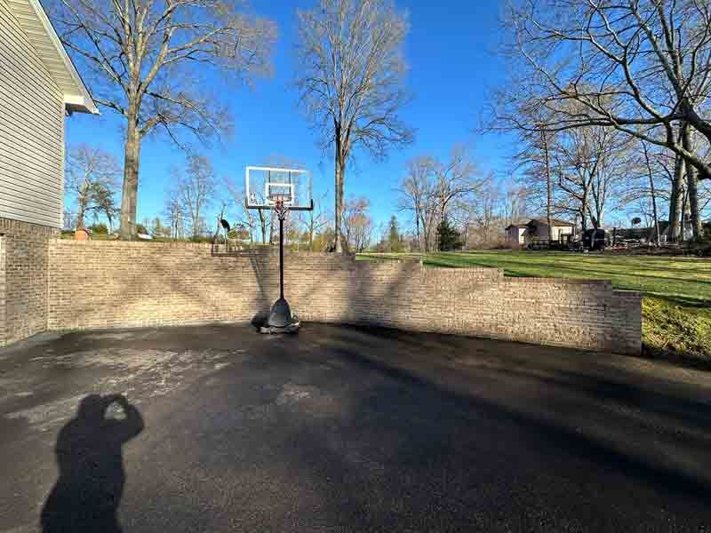 Outdoor basketball hoop on a driveway beside a brick wall, with leafless trees and blue sky.