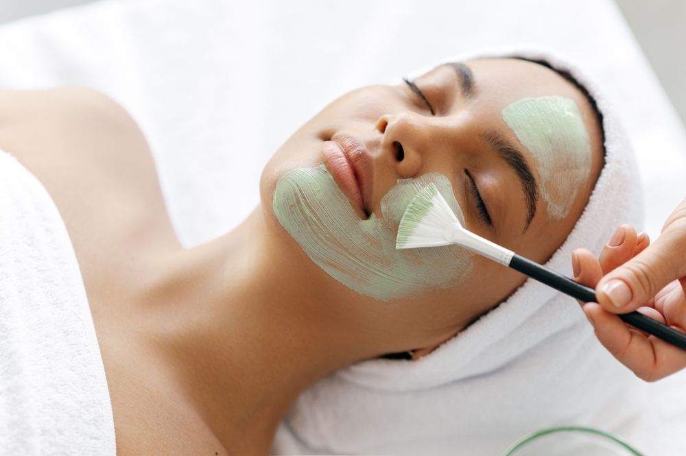 Person receiving a mint-green facial mask application with a brush in a spa setting