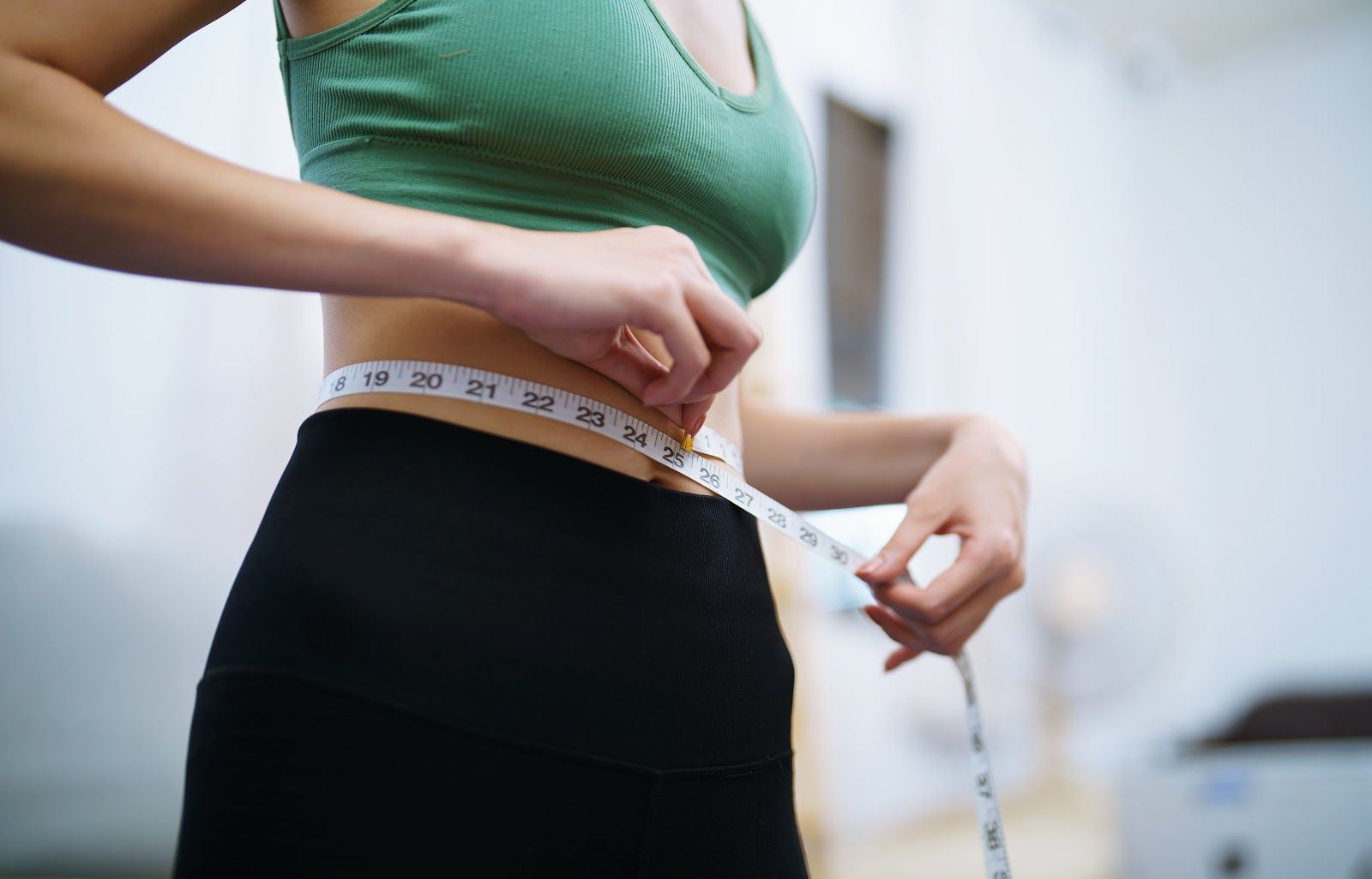 Woman measuring her waist with a tape measure, wearing a green top and black leggings