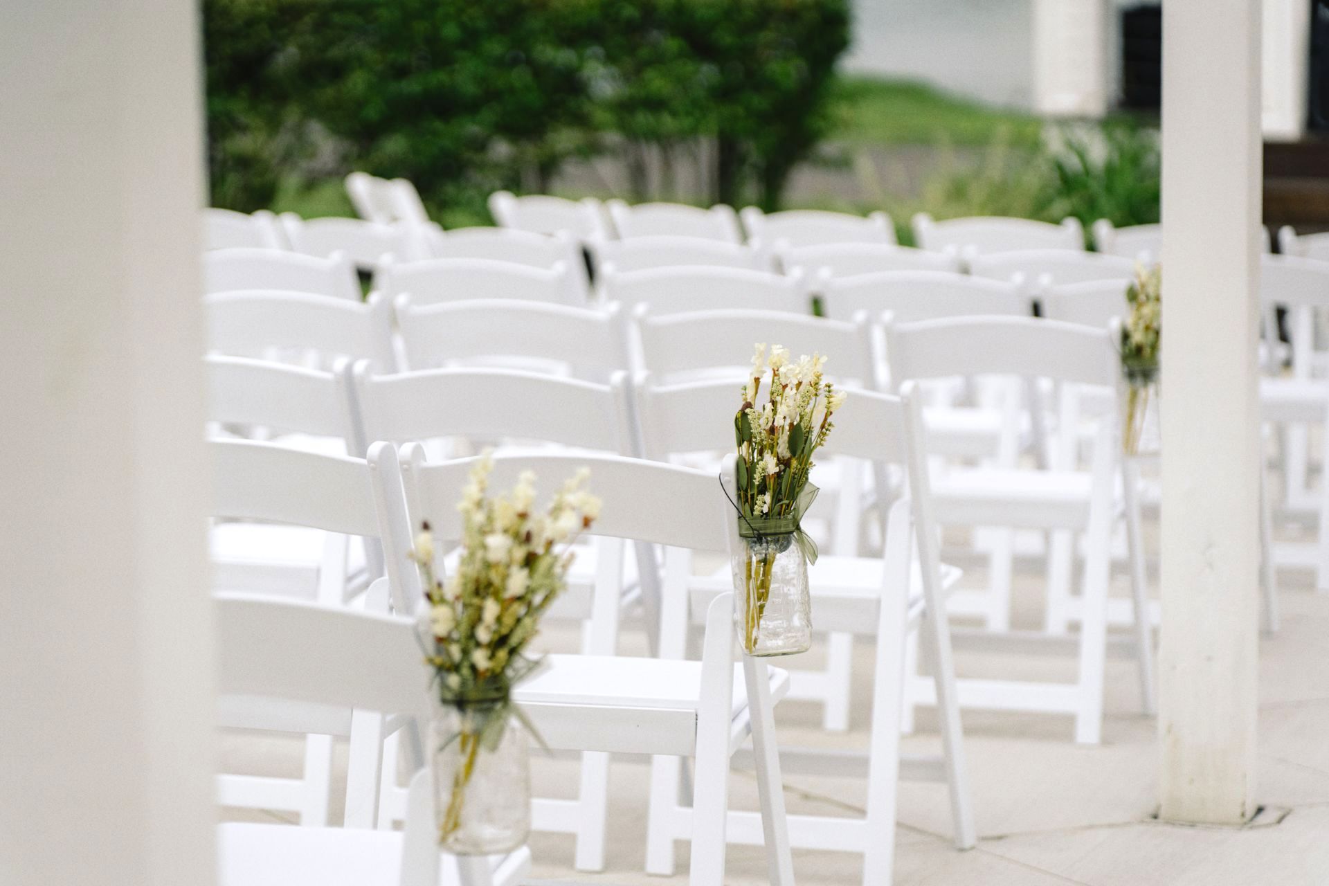 A row of white chairs with vases of flowers on them for a wedding ceremony.