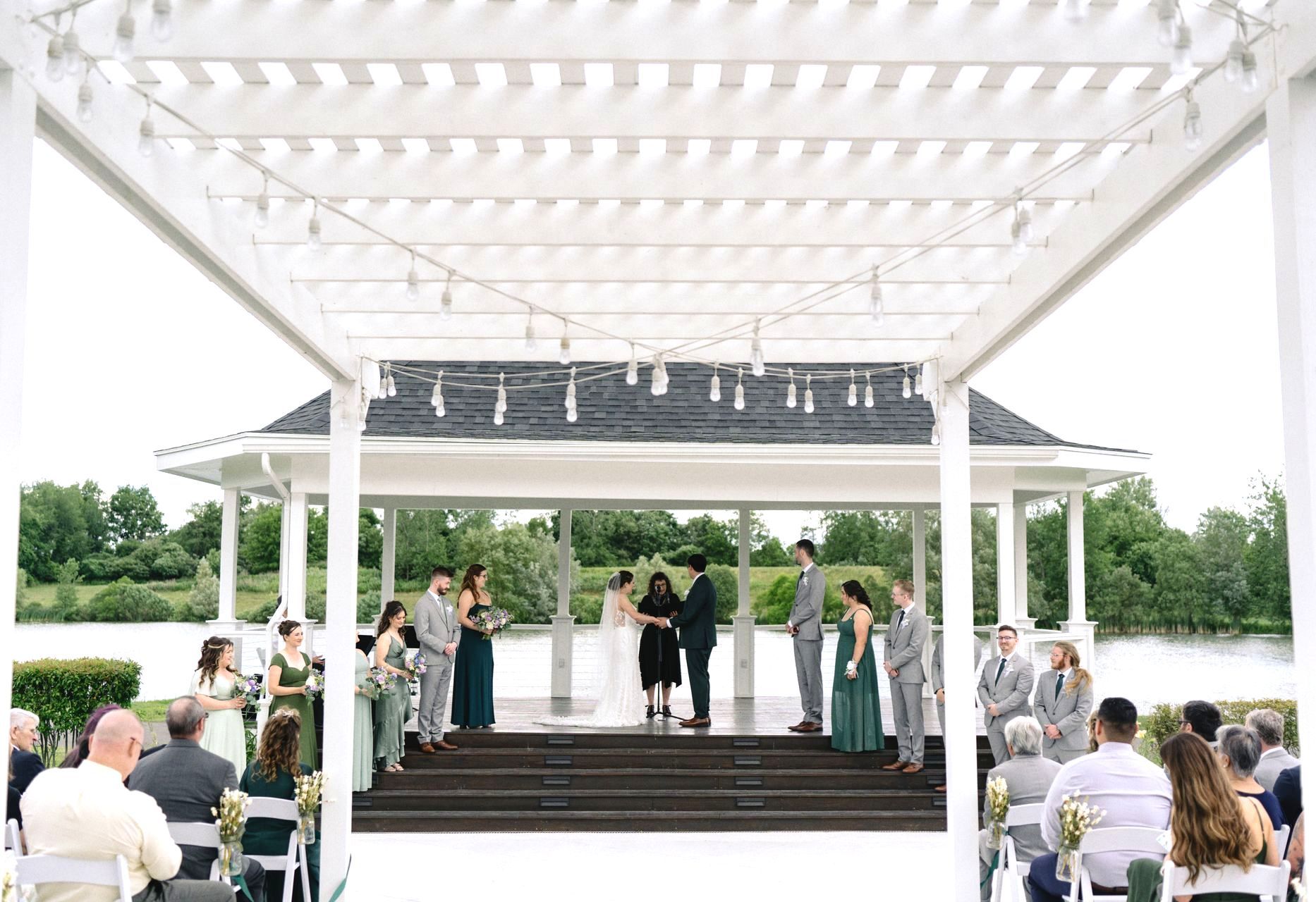 A group of people are sitting under a pergola at a wedding ceremony.