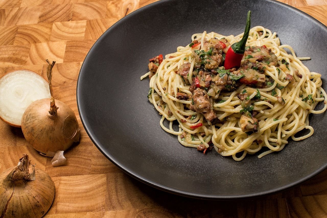 Spaghetti-Gericht mit roter Chili-Garnitur, serviert auf einem schwarzen Teller, dazu Zwiebeln auf einer Holzoberfläche.