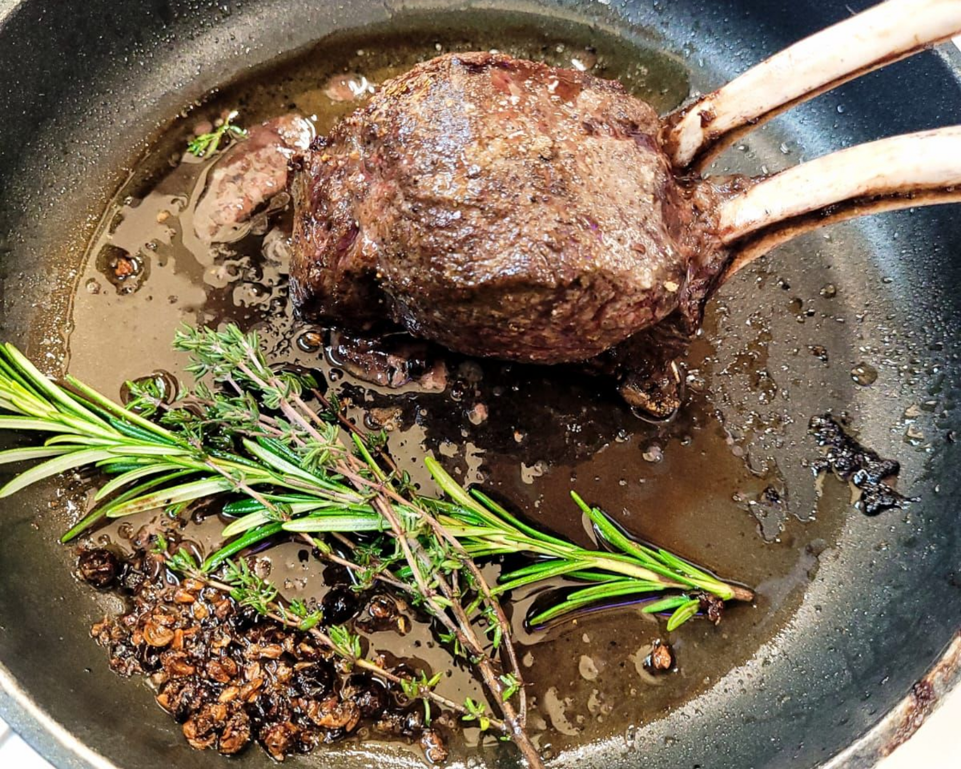 Ein angebratenes Stück Fleisch mit Rippenknochen, Rosmarin und Thymian in der Pfanne.