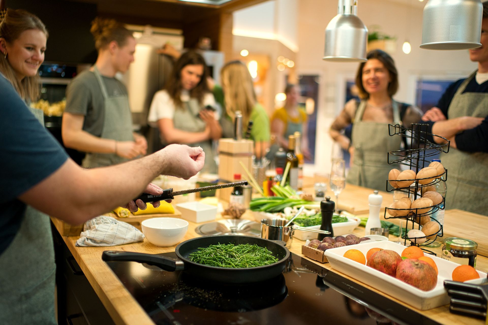 Menschen in einer Küche. Einige kochen, andere schauen zu. Lebensmittel und Kochutensilien auf einer Arbeitsplatte.