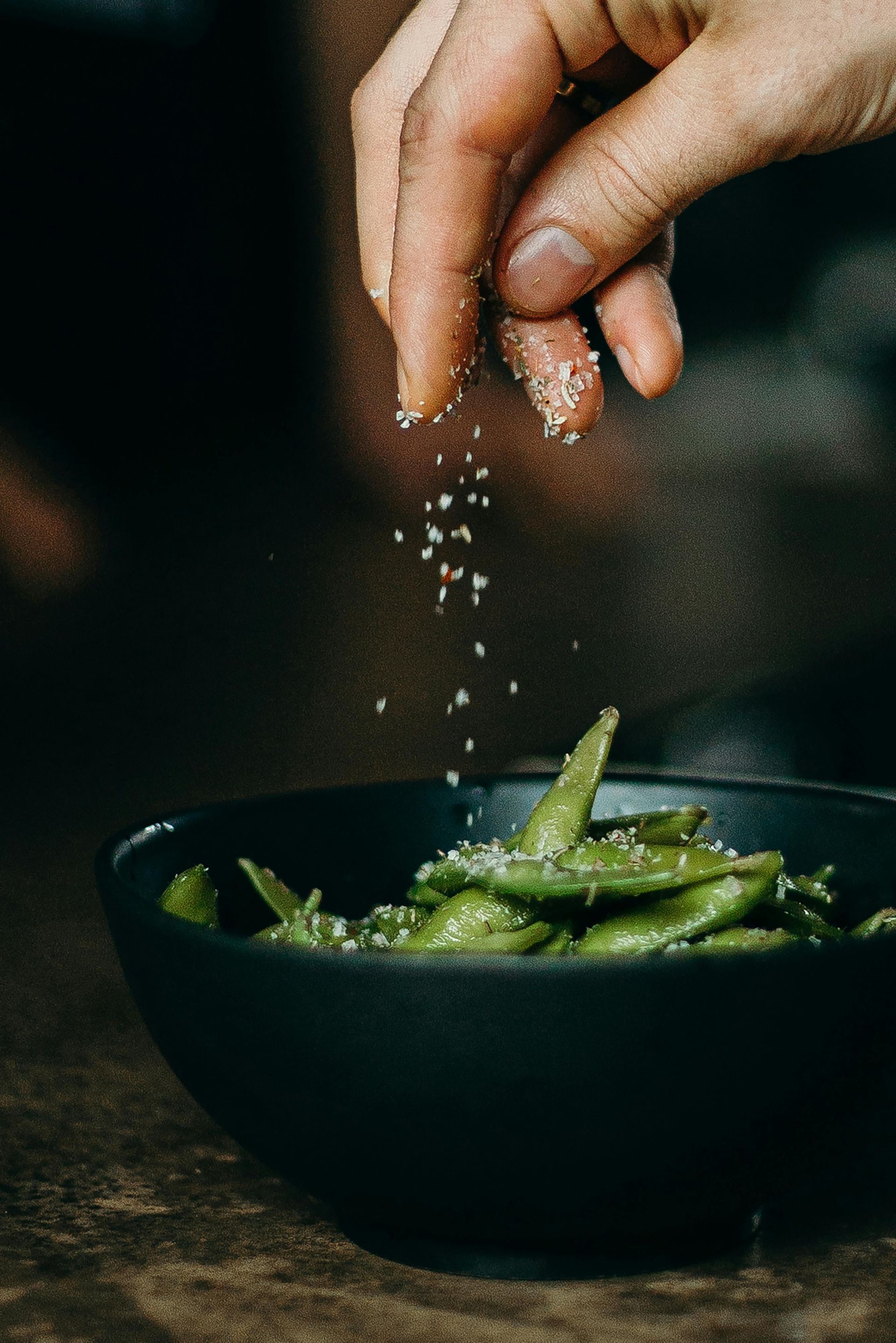 Streuen Sie mit der Hand Salz über eine Schüssel mit grünen Edamame. Dunkle Schüssel, dunkles Gedeck.