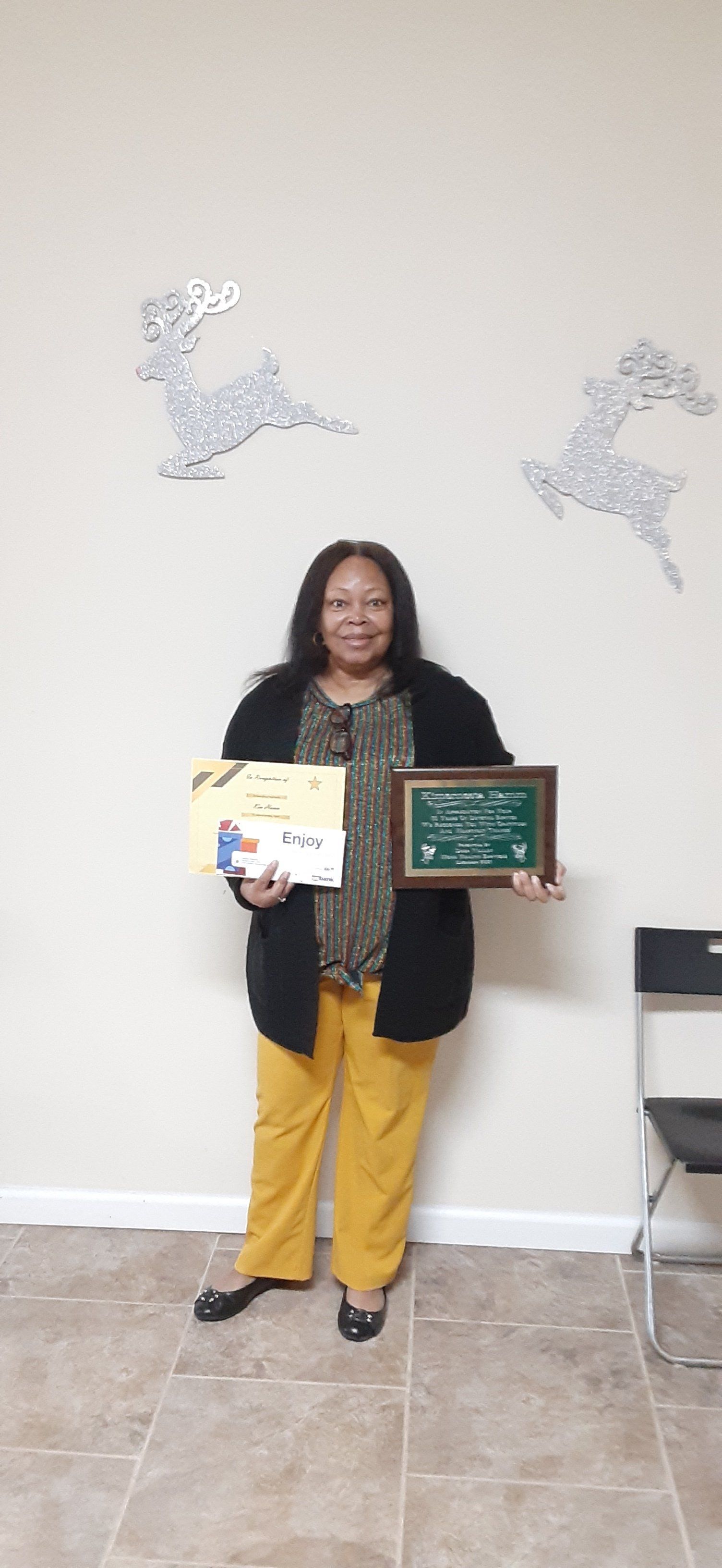 A smiling woman stands holding certificates; wall with reindeer decor behind her. She wears a black sweater, mustard pants.