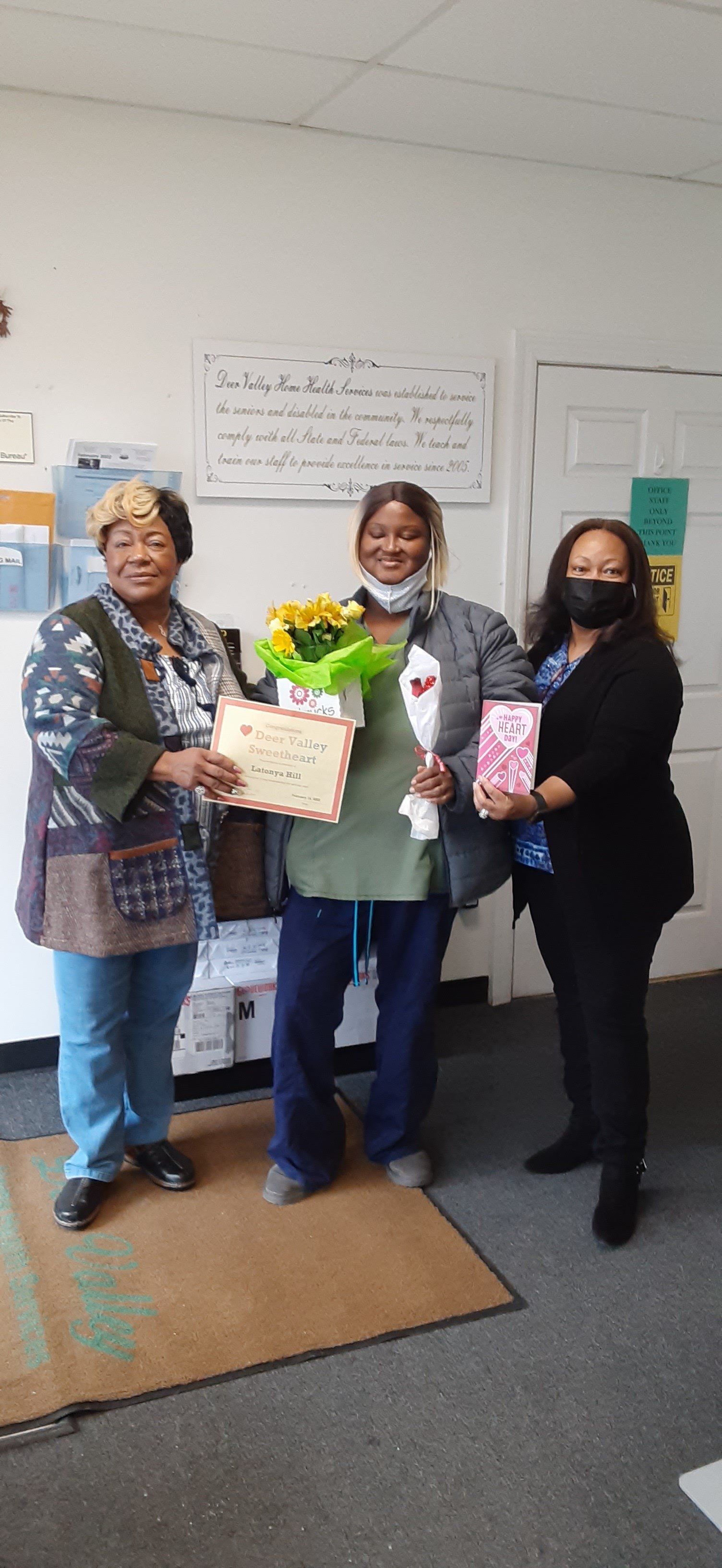 Three women smiling, one holding flowers, another a gift, indoors near a whiteboard and door.