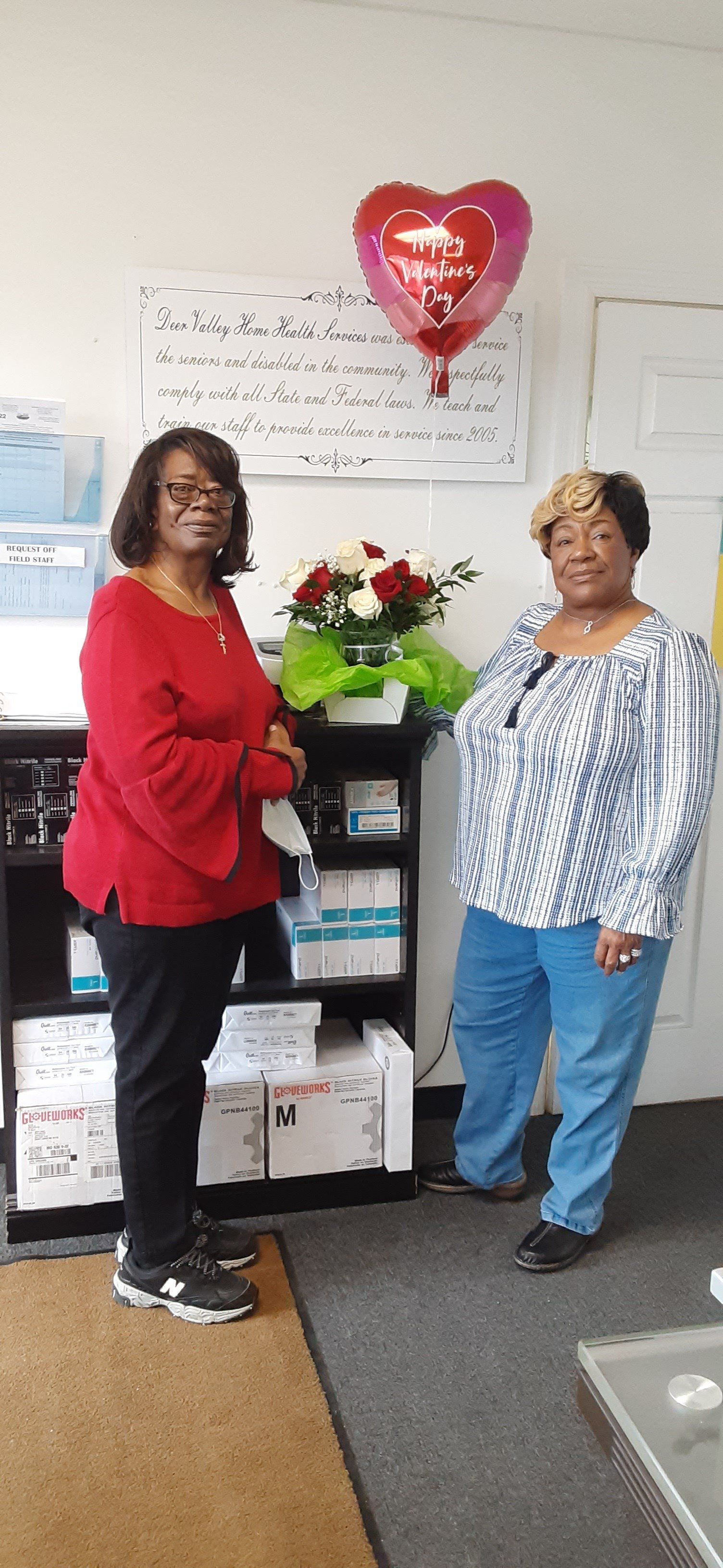 Two women stand near a shelf with flowers and a heart balloon indoors. One wears red, the other blue.