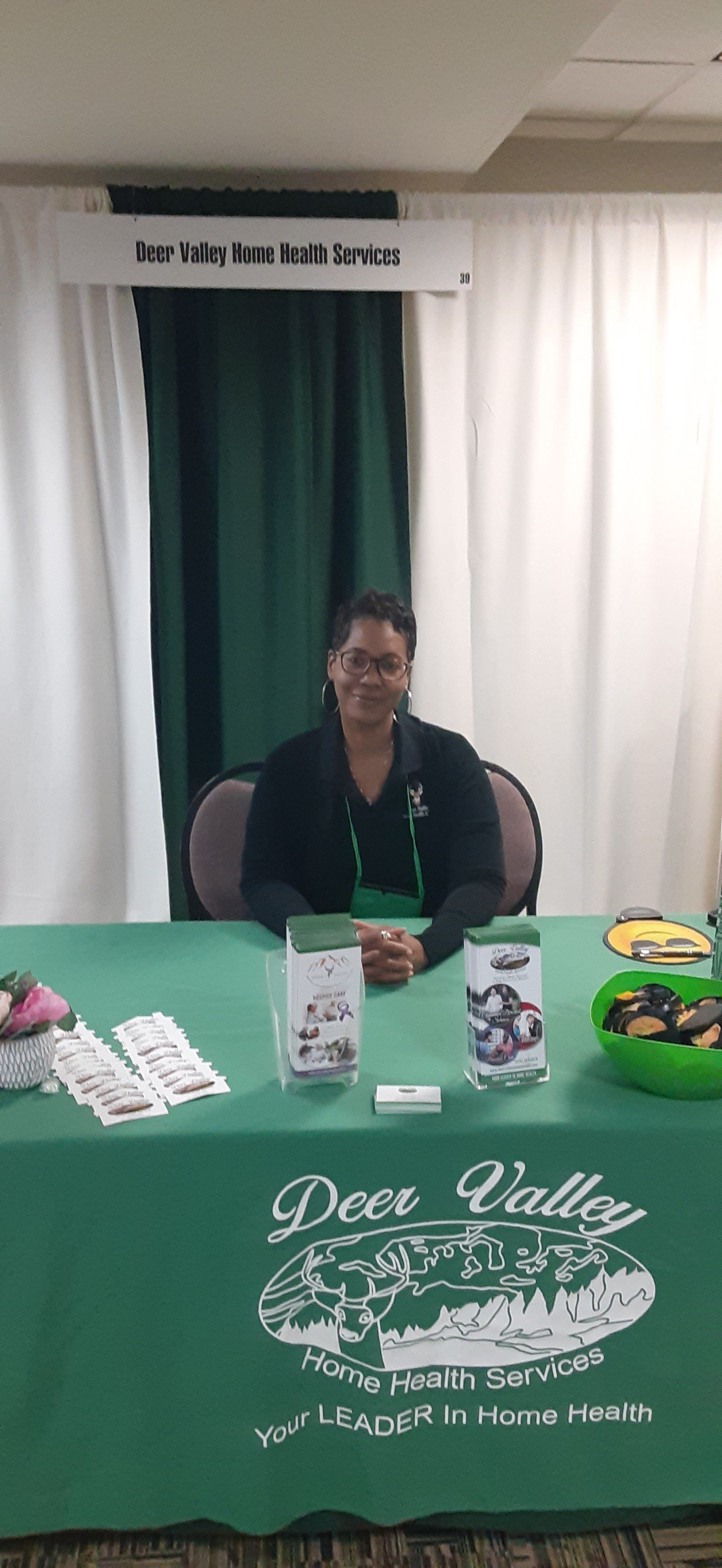 Woman at a table with a Deer Valley Farms logo. She sits smiling, in front of a green table and green curtains.