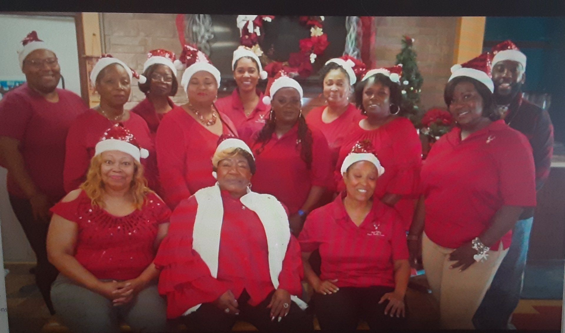 Group of people in red outfits and Santa hats, smiling, posing indoors in front of Christmas decorations.