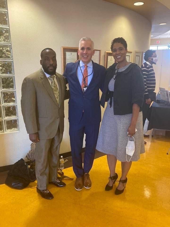 Three people in suits pose indoors, smiling. Two men and one woman stand next to each other. Bright yellow floor.