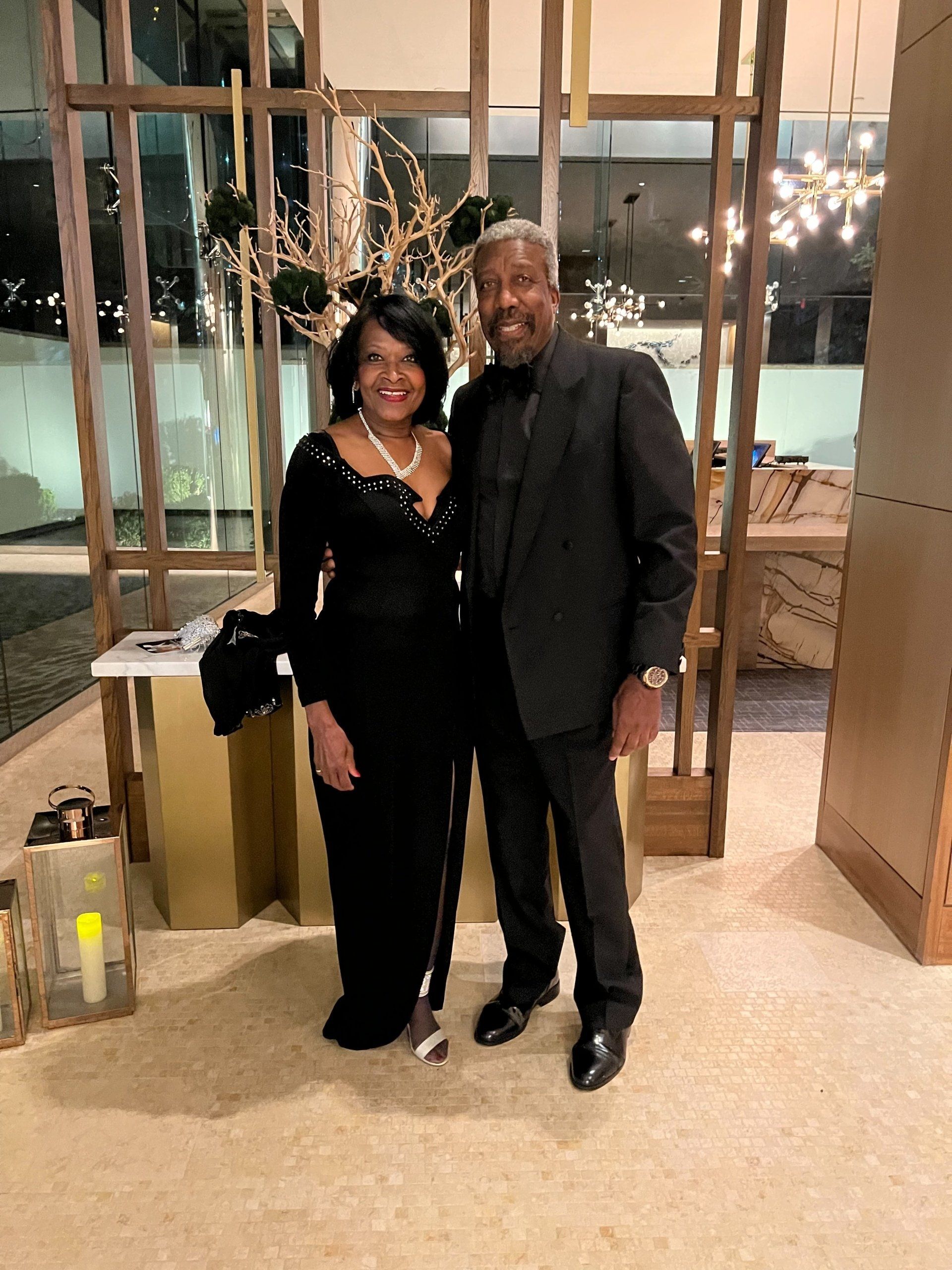 An elegant Black couple poses. Woman in black gown and pearls with cane, man in suit. Interior, neutral tones.