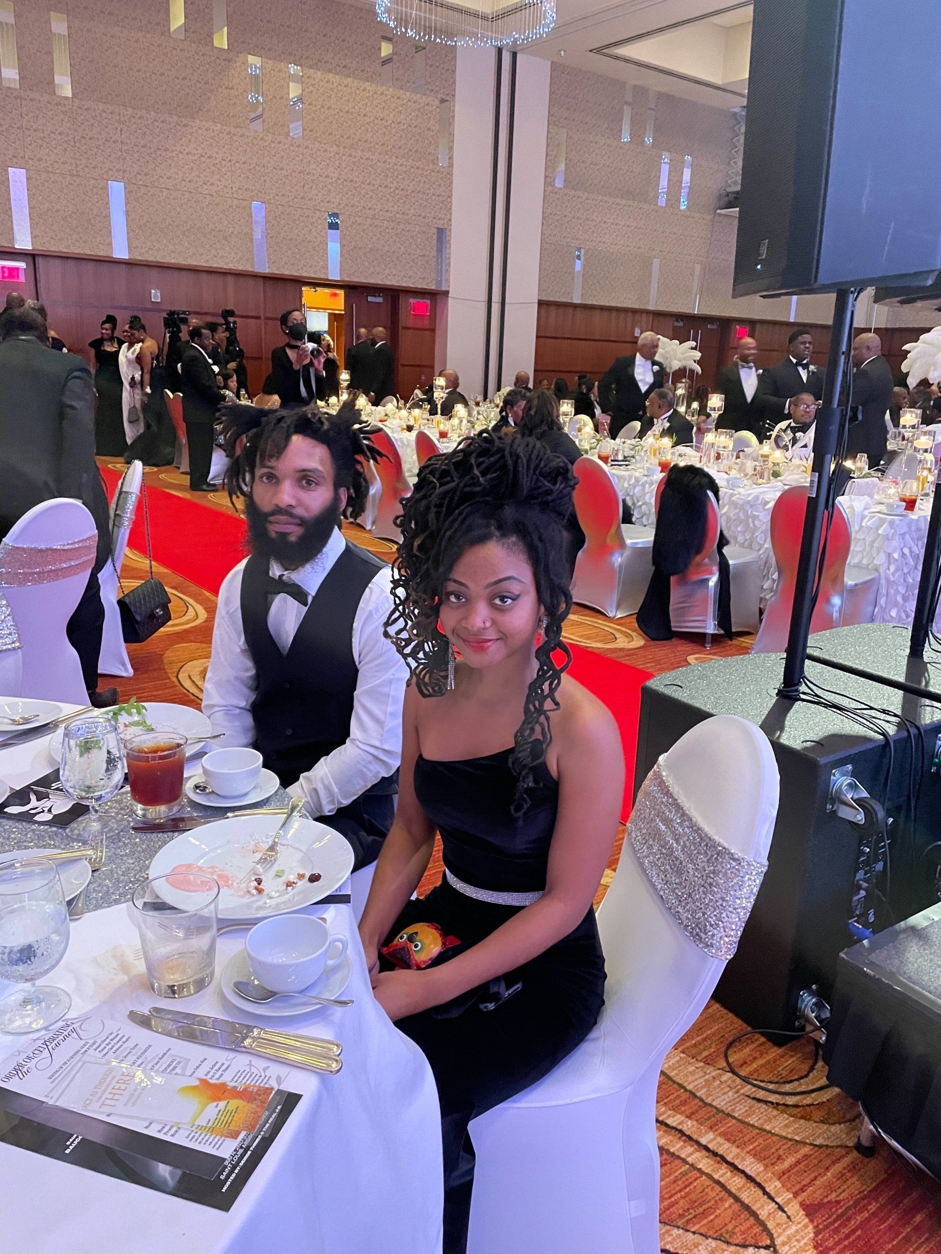 A formally dressed couple sits at a table in a ballroom. She wears a black dress, he wears a vest.