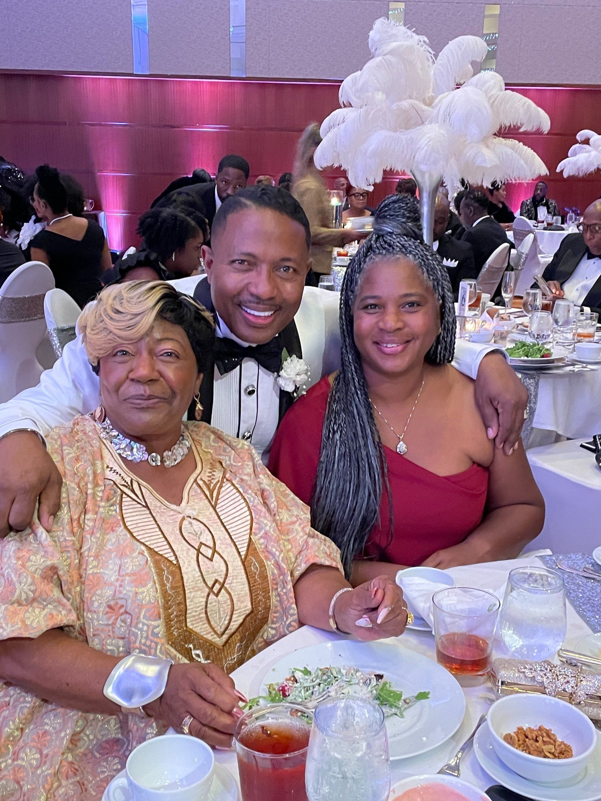 A man in a tuxedo smiles, embracing two women at a formal event. White centerpieces.