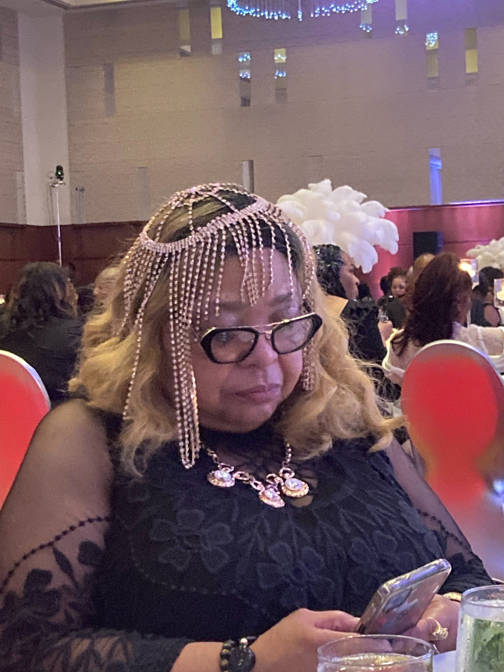 Woman at a formal event wearing a jeweled headpiece and holding a phone, seated at a table.