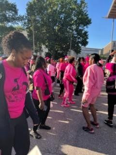 Group of people in pink clothing dancing outdoors under a blue sky, likely at a breast cancer awareness event.