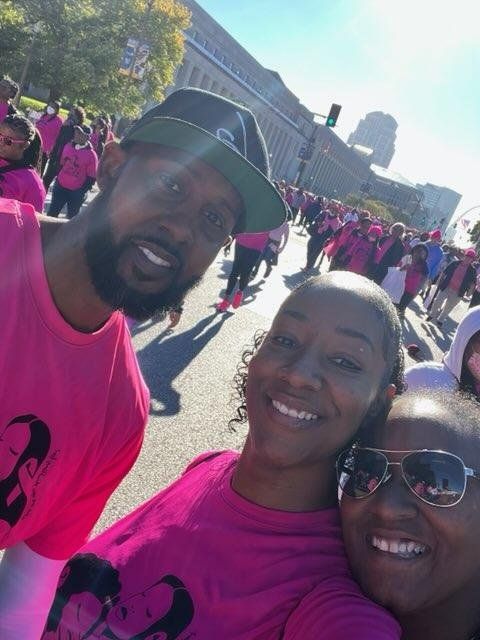 Three people smiling, wearing pink shirts, participating in a breast cancer awareness walk. City street setting.