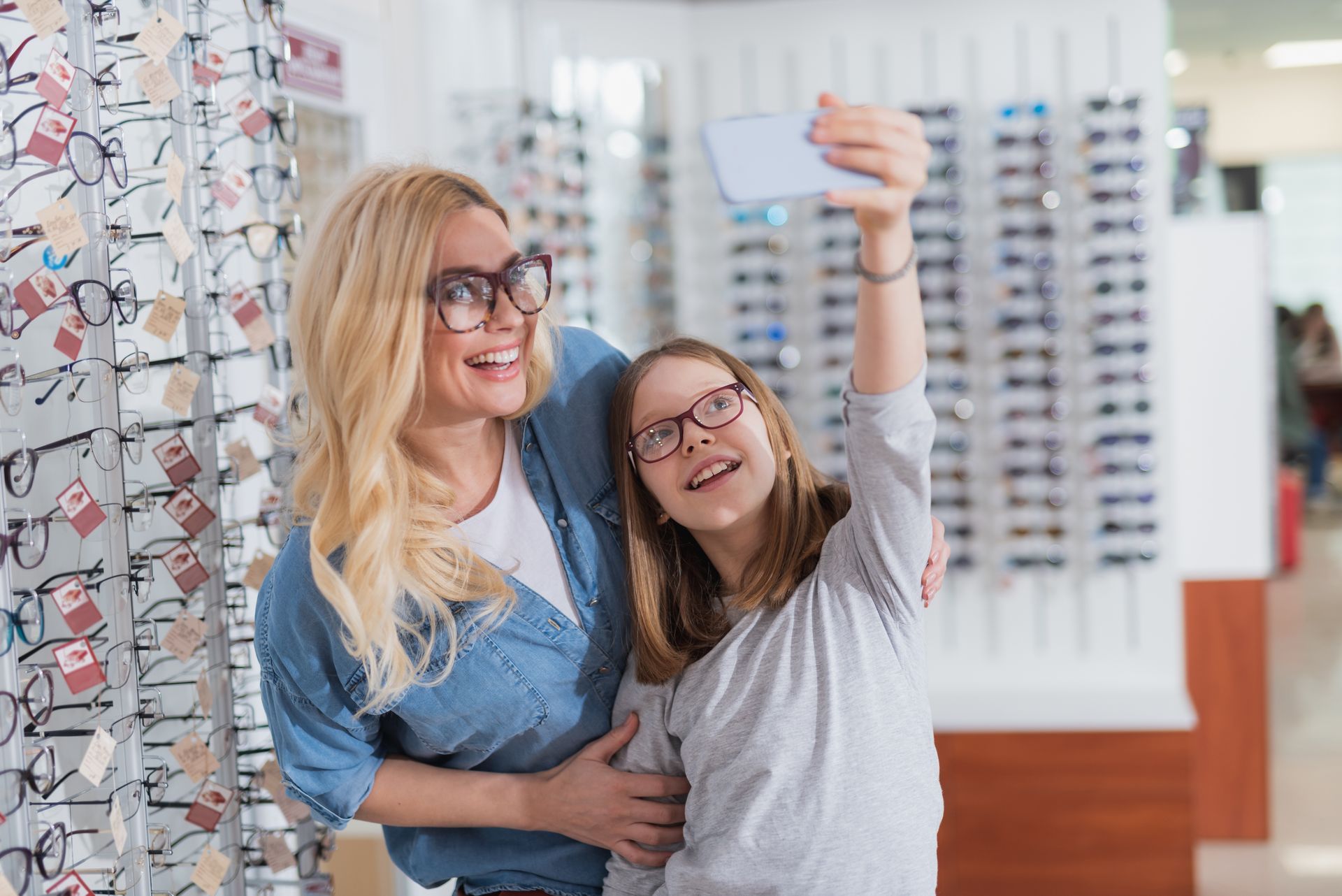 Two smiling people taking a selfie in an eyeglasses store, with glasses displays in the background